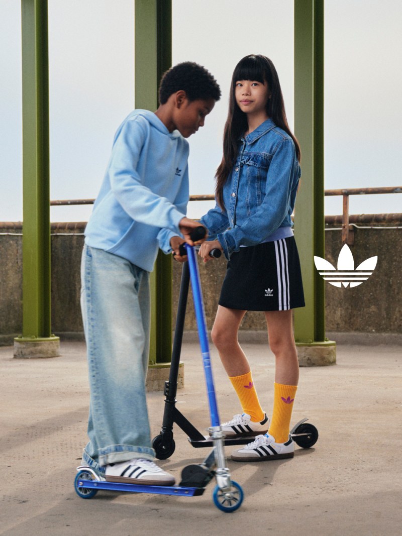 A girl and boy wear white adidas Samba shoes while posing with a scooter on a concrete street.  
