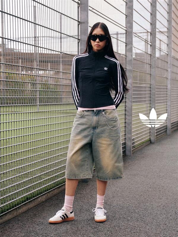A young woman wearing a black Adidas top and light-colored denim shorts stands in front of a metal fence in an urban setting.