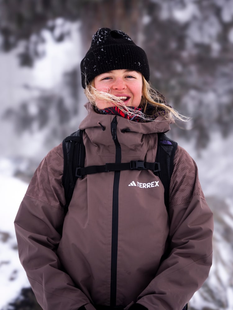 A person wearing a warm winter jacket and hat is standing in a snowy, forested environment.
