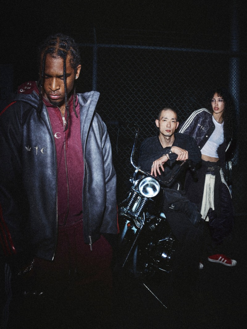 The image shows a man in a red jacket standing in the foreground, with a woman sitting on a motorcycle in the background and another woman standing next to her.