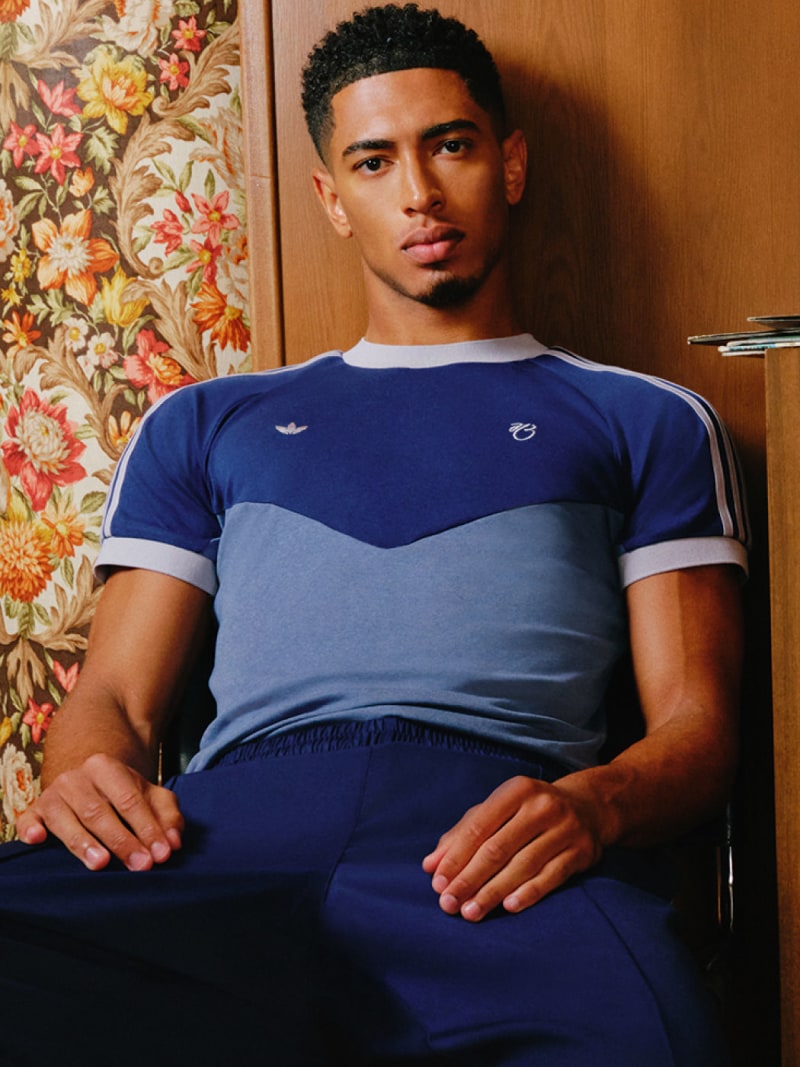 Jude Bellingham leans casually against a wall, fully dressed in the adidas Bellingham collection, looking straight into the camera.