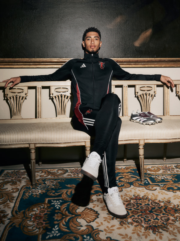 Jude Bellingham wearing a black and red athletic outfit is seated on an ornate, upholstered bench against a backdrop of intricate, patterned carpeting and decorative architectural elements.