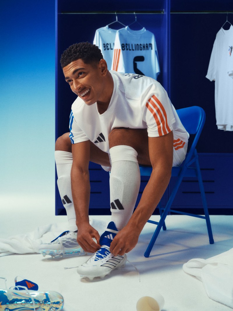 Jude Bellingham ata sus botas adidas Predator rojas, blancas y azules mientras está sentado con su equipación completa sobre una silla azul.