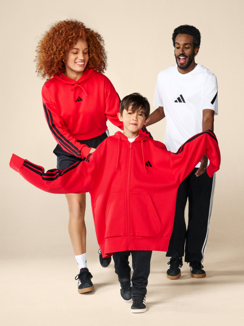 A woman and man in adidas clothing laugh as they help a young boy put on an oversized red adidas hoodie.