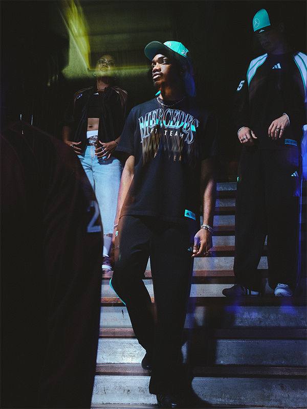 The image shows a group of young people, likely in their late teens or early 20s, standing on a set of stairs in a dimly lit environment. The individuals are wearing casual apparel, including a t-shirt with a graphic design, and appear to be engaged in some kind of social activity or gathering.