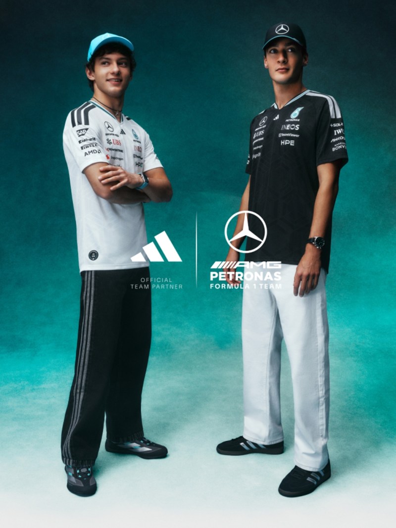 Two individuals wearing Mercedes Formula 1 team uniforms stand in front of a teal background with the Mercedes logo prominently displayed.
