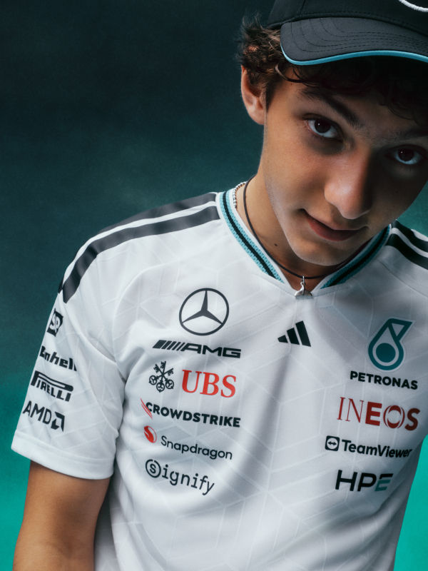 A young man wearing a white sports jersey with various sponsor logos, standing against a dark green background.