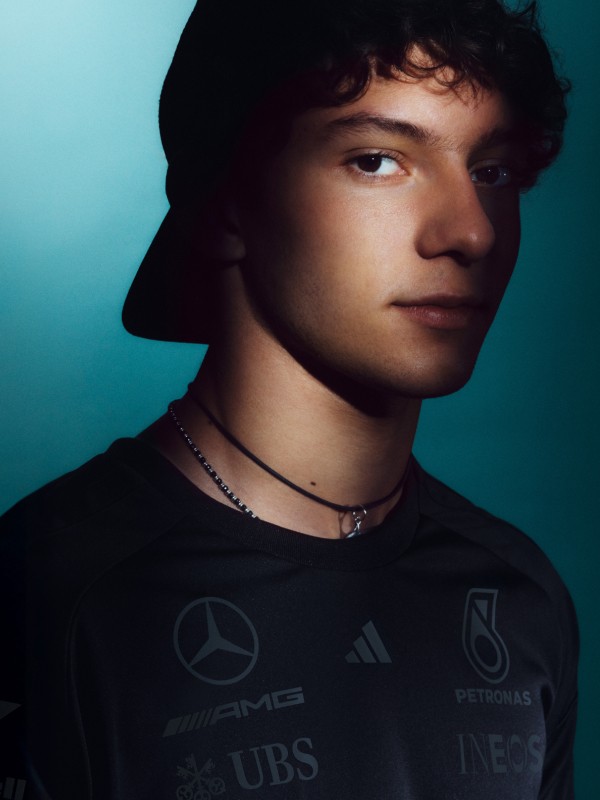 A young man with dark, curly hair and a serious expression, wearing a black shirt with logos, set against a teal-coloured background.
