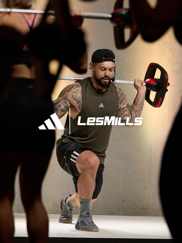 A muscular man with tattoos and a beard is performing a workout exercise while wearing Lesmills branded athletic gear, with a dark background and other people visible in the background.