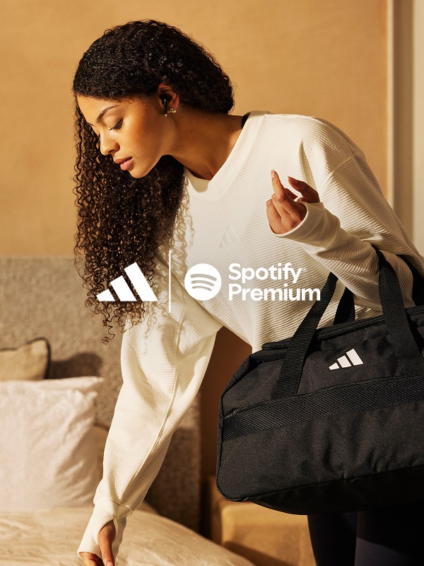Eine junge Frau mit lockigem, dunklem Haar in einem weißen Spotify Premium Sweatshirt sitzt in einer schwarzen Sporttasche auf einem Bett.