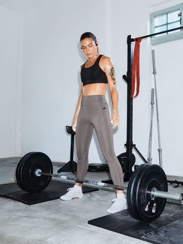 ผู้หญิงในชุดกีฬายืนอยู่ในยิมที่มีอุปกรณ์ออกกำลังกายและบาร์เบลที่มองเห็นได้ในพื้นหลัง