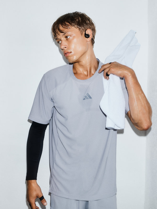 Un joven usando una camisa deportiva gris y shorts negros se para contra un fondo blanco, sosteniendo una toalla blanca.