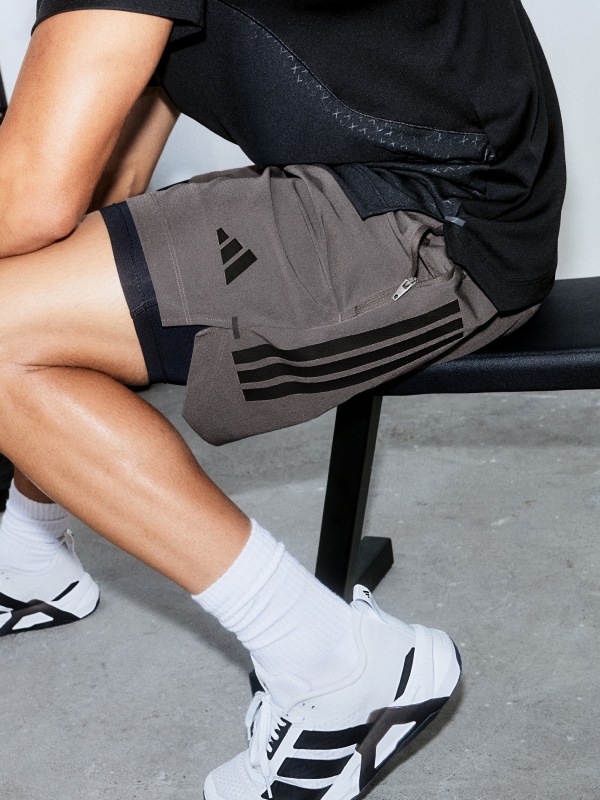 La imagen muestra la parte inferior del cuerpo de una persona usando shorts deportivos grises y zapatillas blancas, sentada en una silla negra sobre un fondo blanco liso.