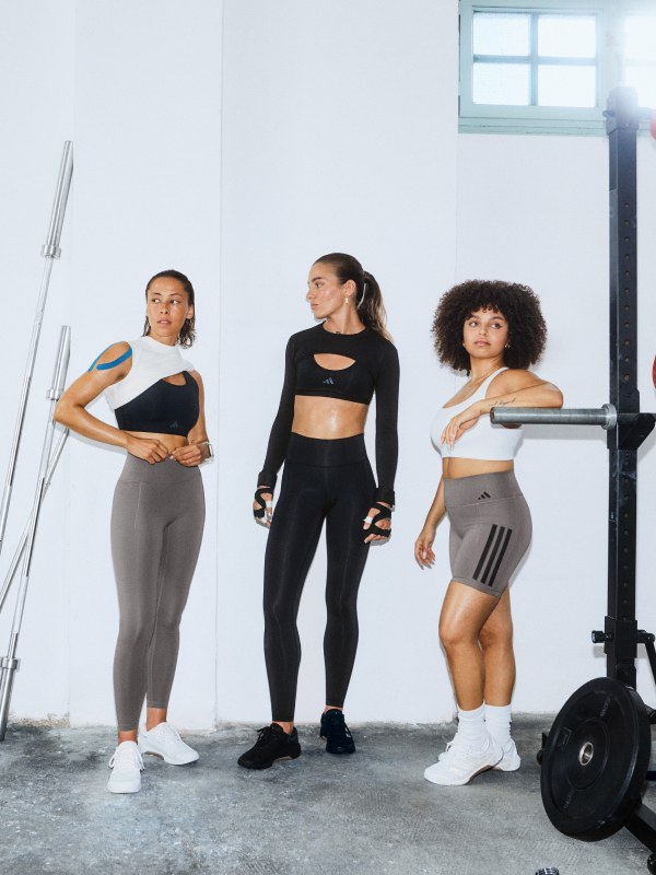 Tres mujeres en ropa deportiva paradas en un gimnasio con equipo para ejercicios visible en el fondo.