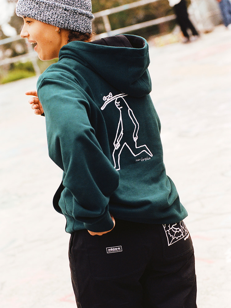 Iemand die een groene hoodie draagt uit de Mark Gonzales Signature Apparel Collection by adidas Skateboarding, met een witte omtrek van het figuurtje met een skateboard op de kop gedrukt op de rug.