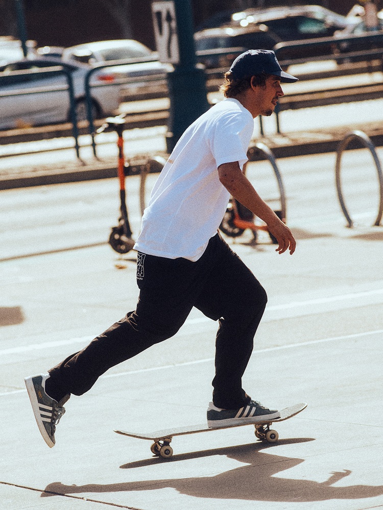 Mężczyzna w białej koszulce i czarnych spodniach jedzie na deskorolce w sneakerach adidas Campus ADV.