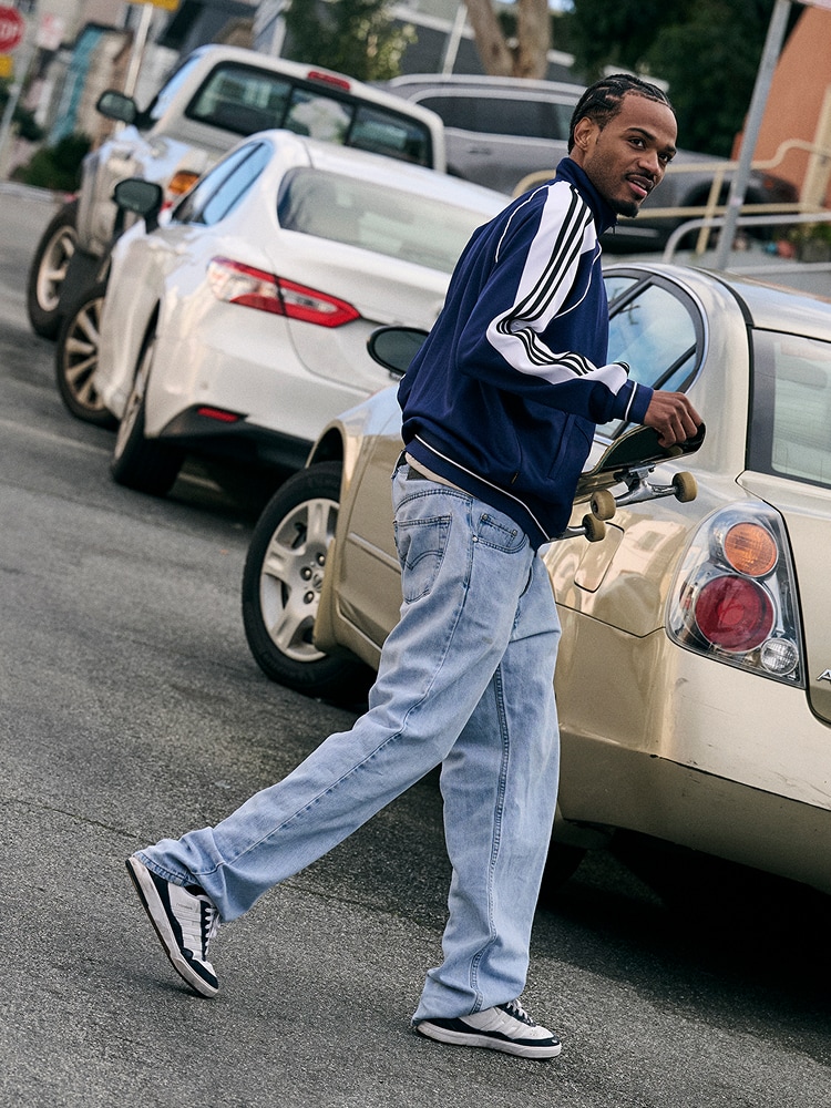 A man in a blue and white striped jacket is standing next to a parked car on a city street, with other vehicles visible in the background.