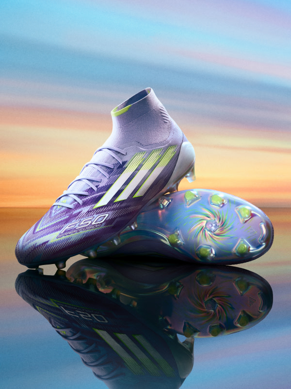 A pair of vibrant purple and green soccer cleats are reflected in a calm, reflective surface against a colorful sunset sky.