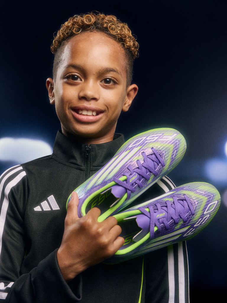 A young, smiling individual wearing a black athletic jacket and holding a pair of vibrant, purple and green athletic shoes.