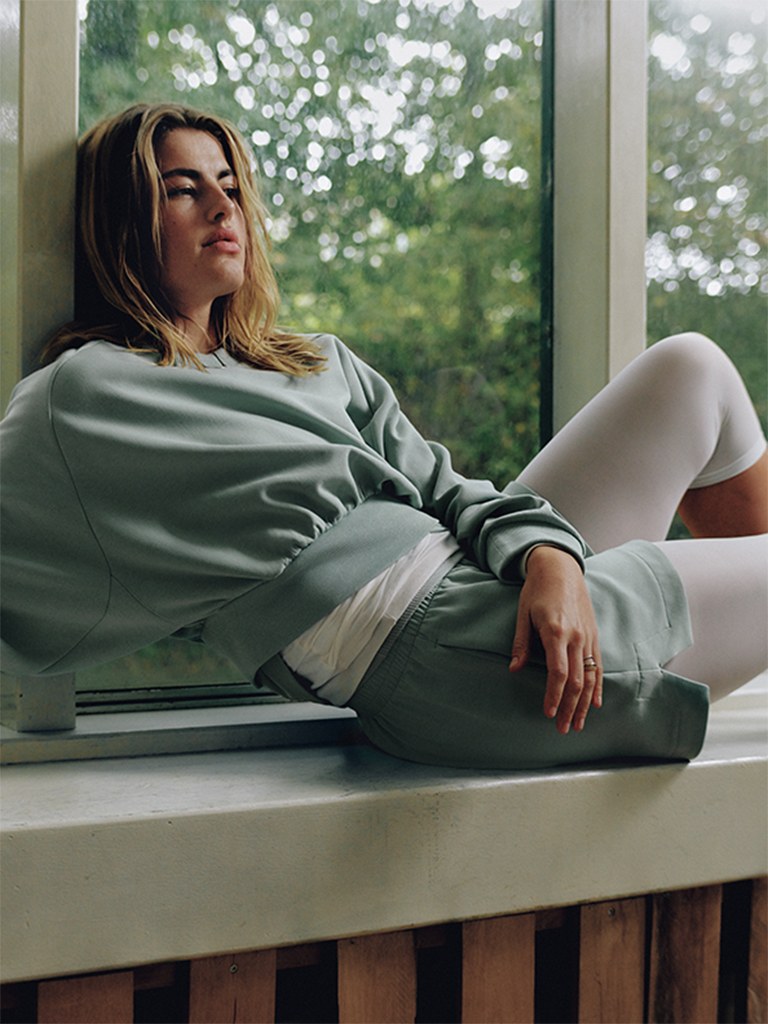 A young woman in a green sweater sits on a windowsill, gazing out at the lush, green foliage visible through the window.