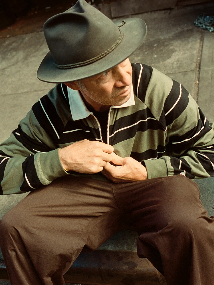 American skateboarder Mark Gonzalez sits on the roadside, fastening the buttons of his adidas x Shmoofoil striped shirt. Wearing a fedora, he gazes off to the right.