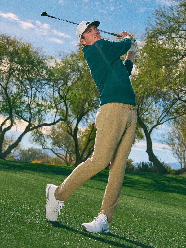 A golfer in a green sweater is swinging a golf club on a grassy golf course surrounded by trees.
