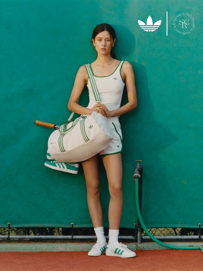 A brown-haired woman leans casually against a green wall, wearing a white tank top, green shorts, and sneakers by adidas x Sporty & Rich.