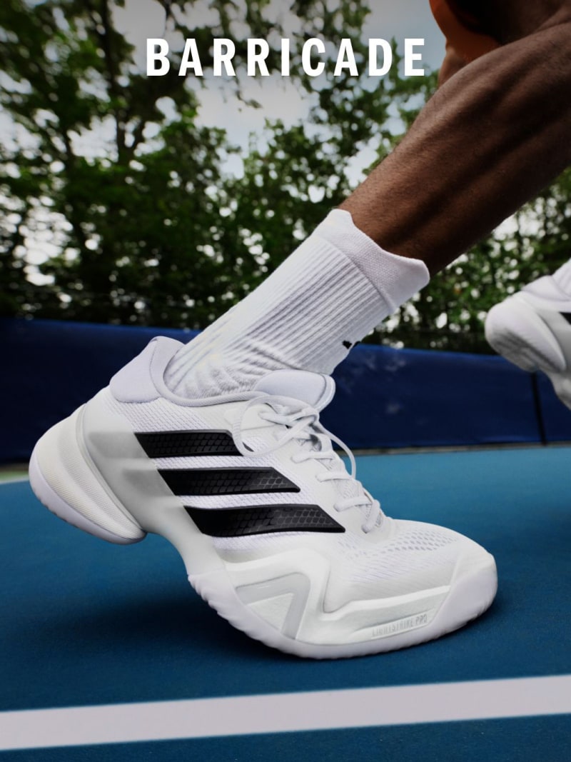 The image shows a pair of white and black athletic shoes with the word "BARRICADE" prominently displayed on the side. The shoes are placed on a blue surface, with a blurred natural background of trees and foliage visible.