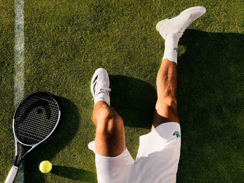 The image shows a person's legs and feet in tennis attire, with a tennis racket and a tennis ball on a grassy tennis court.
