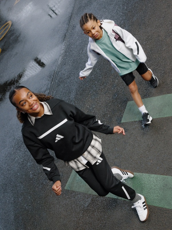 Two young women wearing athletic clothing are standing on a gray floor with a checkered pattern, one appears to be reaching out to the other.