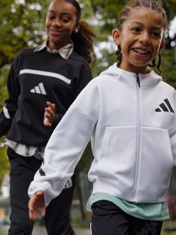 Two young girls, one wearing a black and white athletic jacket and the other wearing a white athletic jacket, are smiling and standing in a park-like setting with trees and greenery in the background.