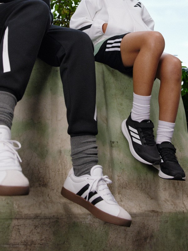 The image shows the lower body of two people sitting on a grassy surface, with one person wearing black pants and the other wearing white pants. The background appears to be a natural setting with some foliage visible.