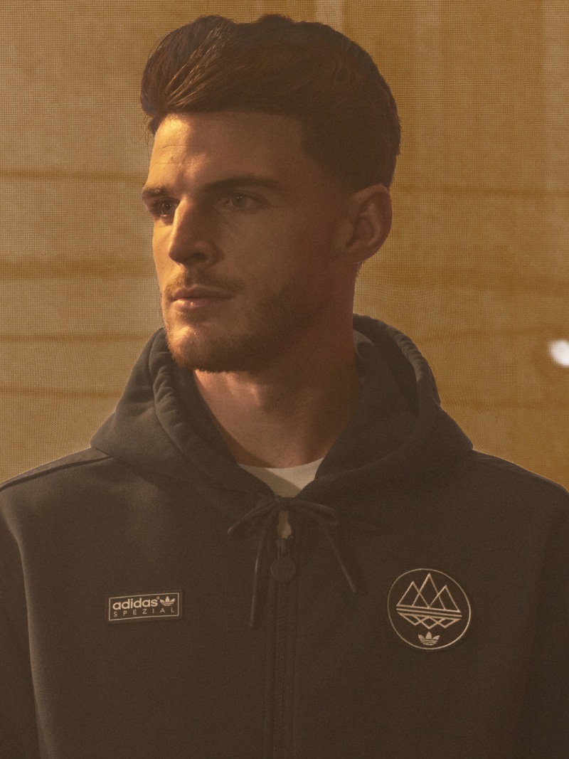 A young man with short dark hair wearing a black jacket with an Adidas logo and a mountain emblem, standing against a plain background.