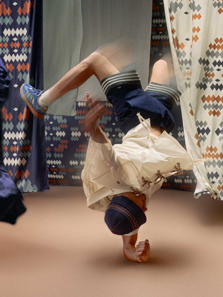 A person in a blue bucket hat and white shirt performs breakdancing with legs extended wearing blue adidas x Edison Chen Silk Gazelle sneakers, surrounded by patterned fabric.