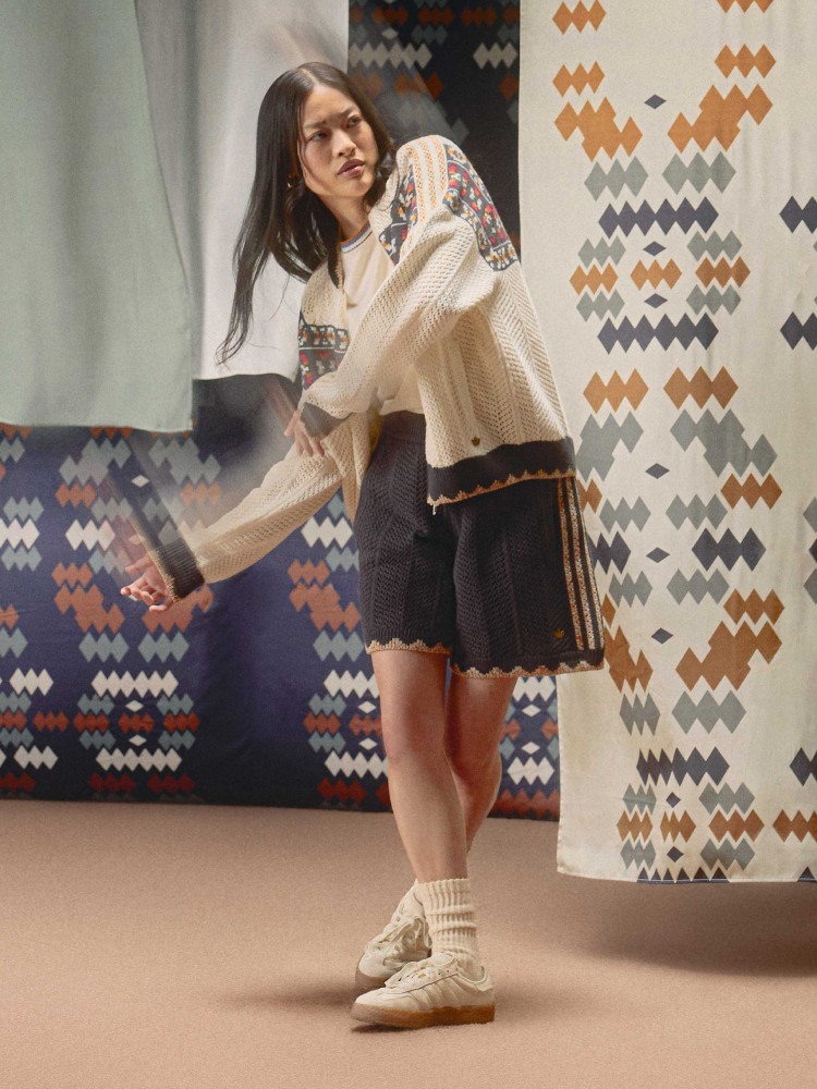 A person in a sweater and skirt wearing the white adidas x Edison Chen Silk Gazelle sneaker stands in a textile-filled room with patterned backgrounds and heavy curtains.