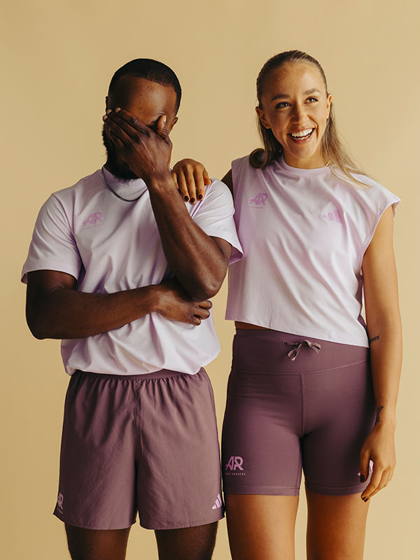 Two people wearing matching pink shirts and shorts are smiling and embracing in front of a plain beige background.
