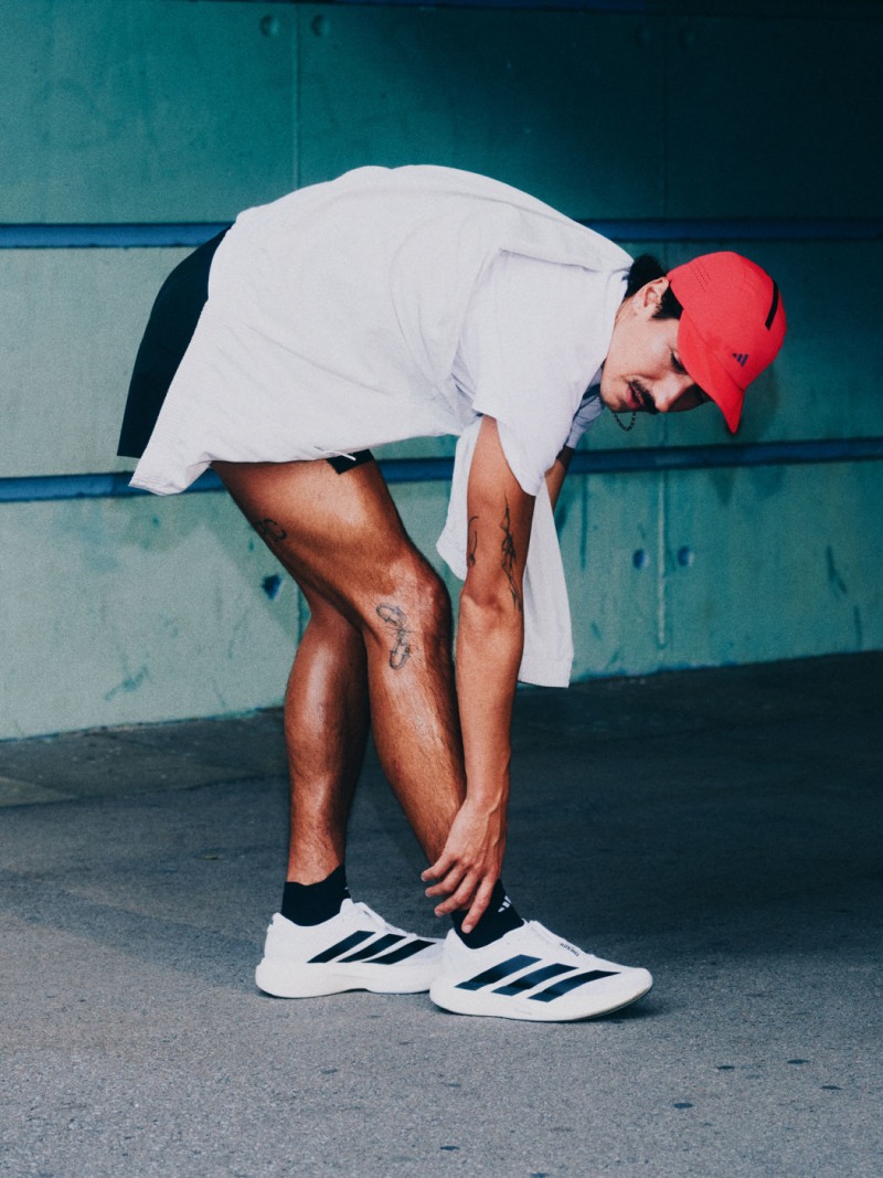 A person in a white outfit and red cap is bending down on a concrete surface with a blue wall in the background.