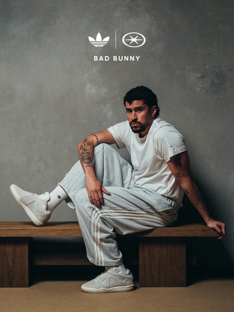 Bad Bunny looks at the camera, sitting on a wooden bench against a textured grey wall, wearing a white T-shirt, light grey adidas track pants, white adidas socks, and white adidas trainers. One leg is bent on the bench, the other foot rests on the floor.