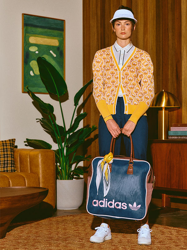 Một người mặc áo thun hoa văn màu vàng, quần jean xanh, và một chiếc mũ trắng đứng trong căn phòng với một cây cối lớn, hình vẽ được đóng khung và một chiếc túi adidas.