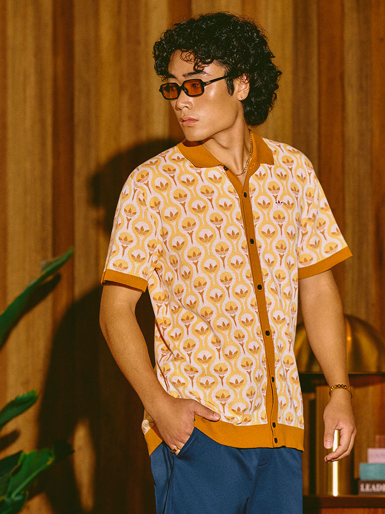 A person with curly hair and glasses wearing a patterned yellow shirt stands in a room with wooden walls and a potted plant in the background.
