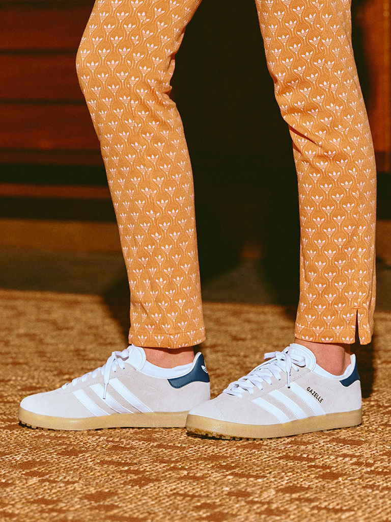 The image shows a person's lower body wearing patterned orange pants and white trainers standing on a wooden floor.