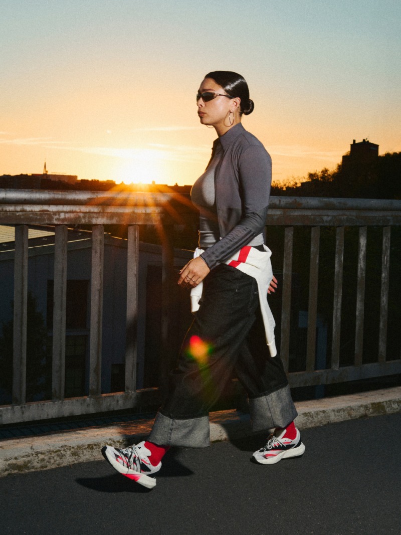Iemand die een grijs shirt, een zwarte broek en rode sneakers draagt, staat op een brug en kijkt bij zonsondergang uit over de skyline van de stad.