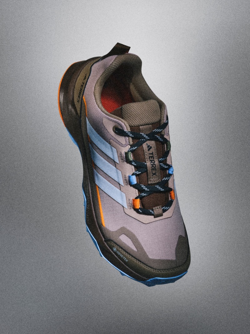 En grå och orange sportsko med snören och en texturerad sula, mot en slätgrå bakgrund.