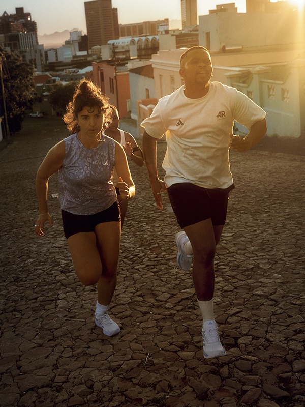 Two people, a man and a woman, are jogging together on a city street with tall buildings in the background, as the sun sets in the distance.