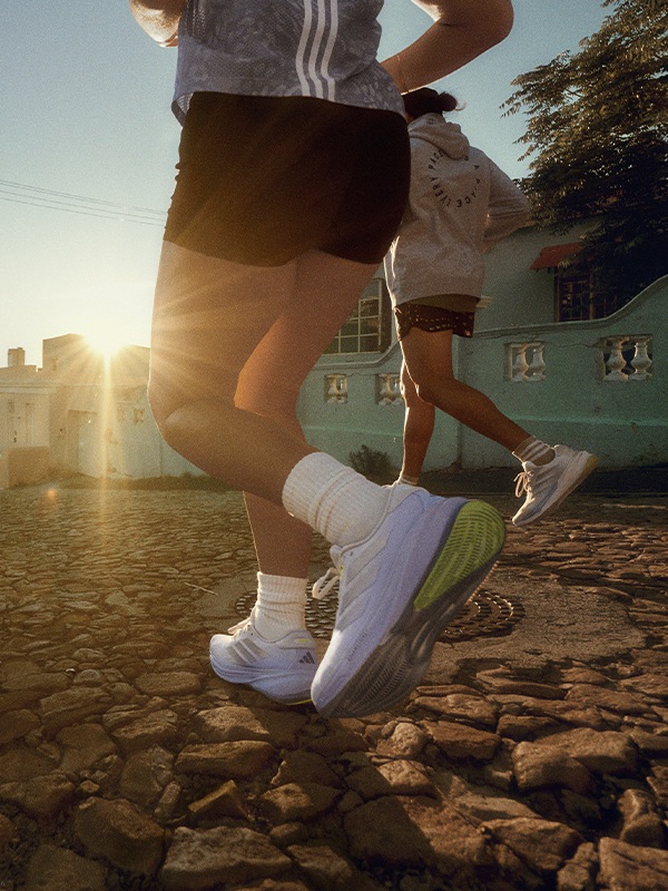A person in athletic clothing is running on a rocky path, with a vibrant green building and bright sunlight in the background.
