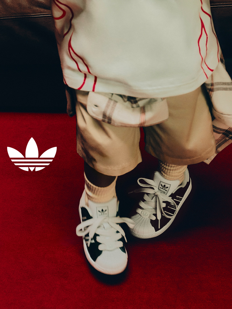 Close-up of a child wearing black-and-white adidas Superstar kids' sneakers on a red carpet, with the Trefoil logo overlayed on the left.