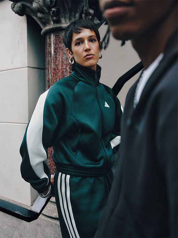 คนสวมชุดแทรคอาดิดาสสีเขียวยืนอยู่ข้างหน้ากำแพงอิฐโดยที่อีกคนหนึ่งมองเห็นได้ในพื้นหลัง