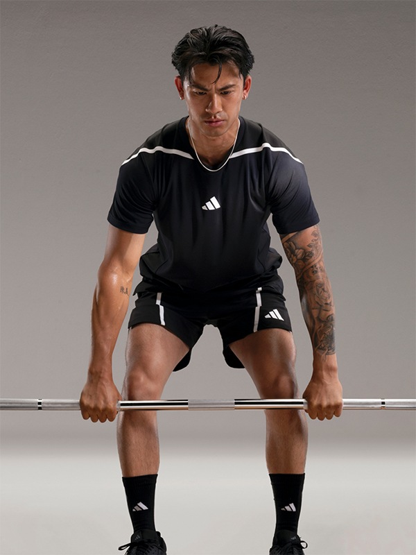 A muscular man in a black athletic shirt and shorts is crouched down, gripping a barbell in front of a plain gray background.
