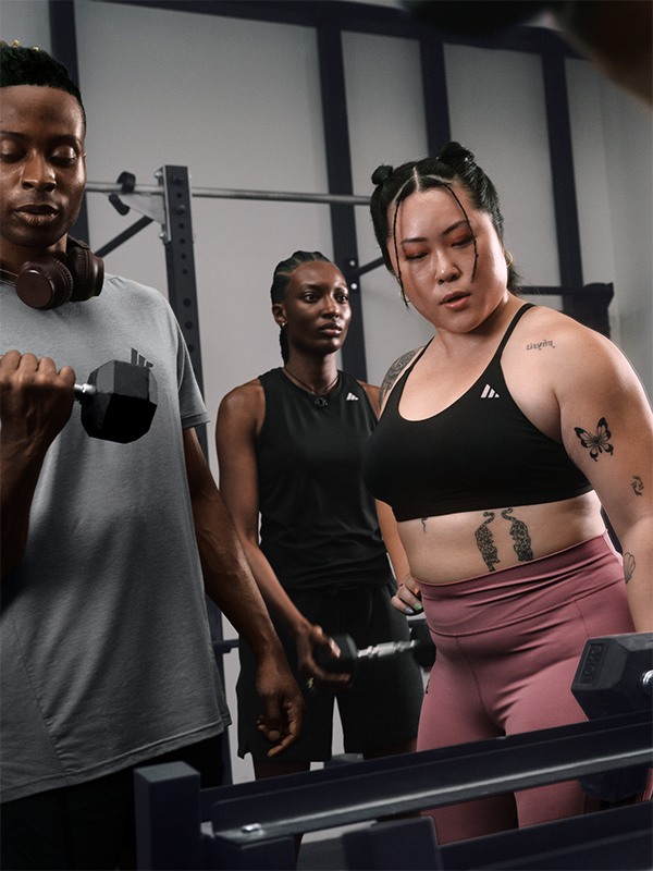 The image shows three individuals, two men and one woman, standing in a gym setting with exercise equipment visible in the background.