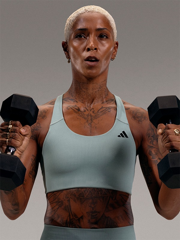 A woman with a shaved head and tattoos on her upper body is holding two dumbbells, standing against a plain gray background.